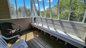 A covered outdoor seating area with a bench and chairs, wooden flooring, and large windows offering a view of trees. - Fair Winds - Oceanside home w/Private Pool - Dogs Allowed! (Southern Shores)