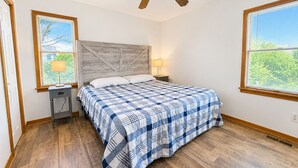 A bedroom with a wooden floor, a bed with a blue and white checkered bedspread, a wooden headboard, two bedside lamps, and a ceiling fan. - “Hank's Hideaway” is a classic, coastal cottage, located just 341 yards to the beach! (Duck)