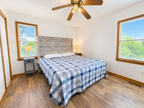 A bedroom with a wooden floor, a bed with a blue and white checkered bedspread, a wooden headboard, two bedside lamps, and a ceiling fan.