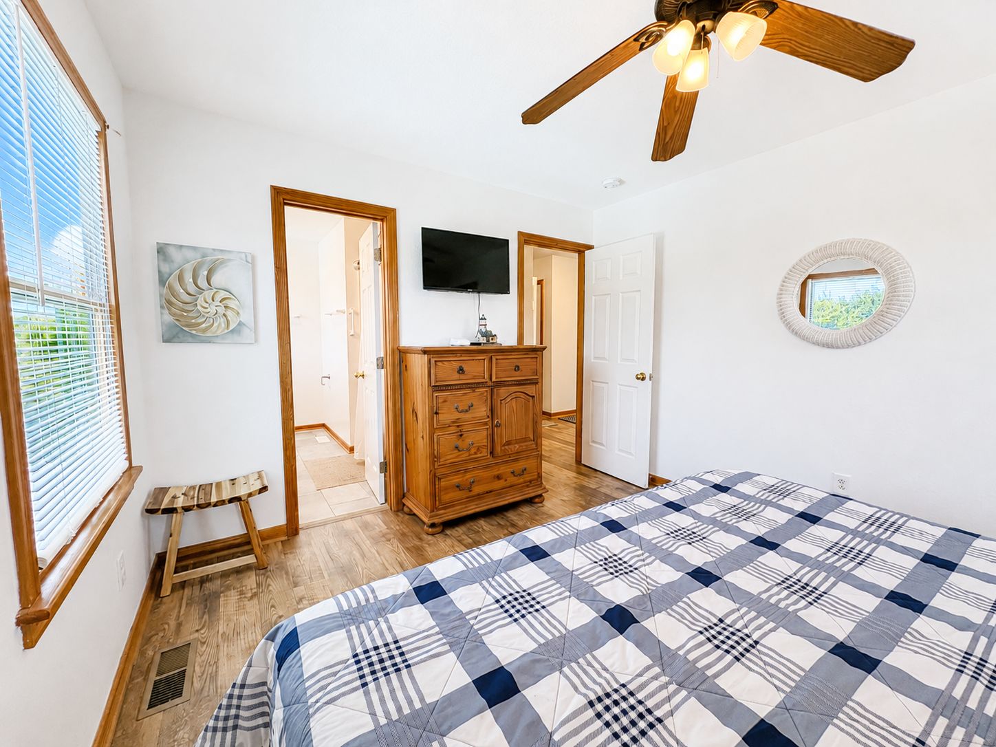 A bedroom with a bed, a wooden dresser, a ceiling fan, a television, and a door leading to another room.