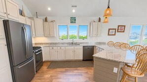A modern kitchen with white cabinets, a stainless steel refrigerator, a built-in oven, and a granite countertop. - “Hank's Hideaway” is a classic, coastal cottage, located just 341 yards to the beach! (Duck)