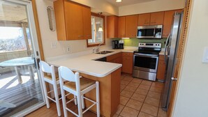 A modern kitchen with wooden cabinets, a white countertop, stainless steel appliances, and a glass door leading to an outdoor area. - Knock On Wood - Directly across from the beach access with terrific ocean views (Duck)