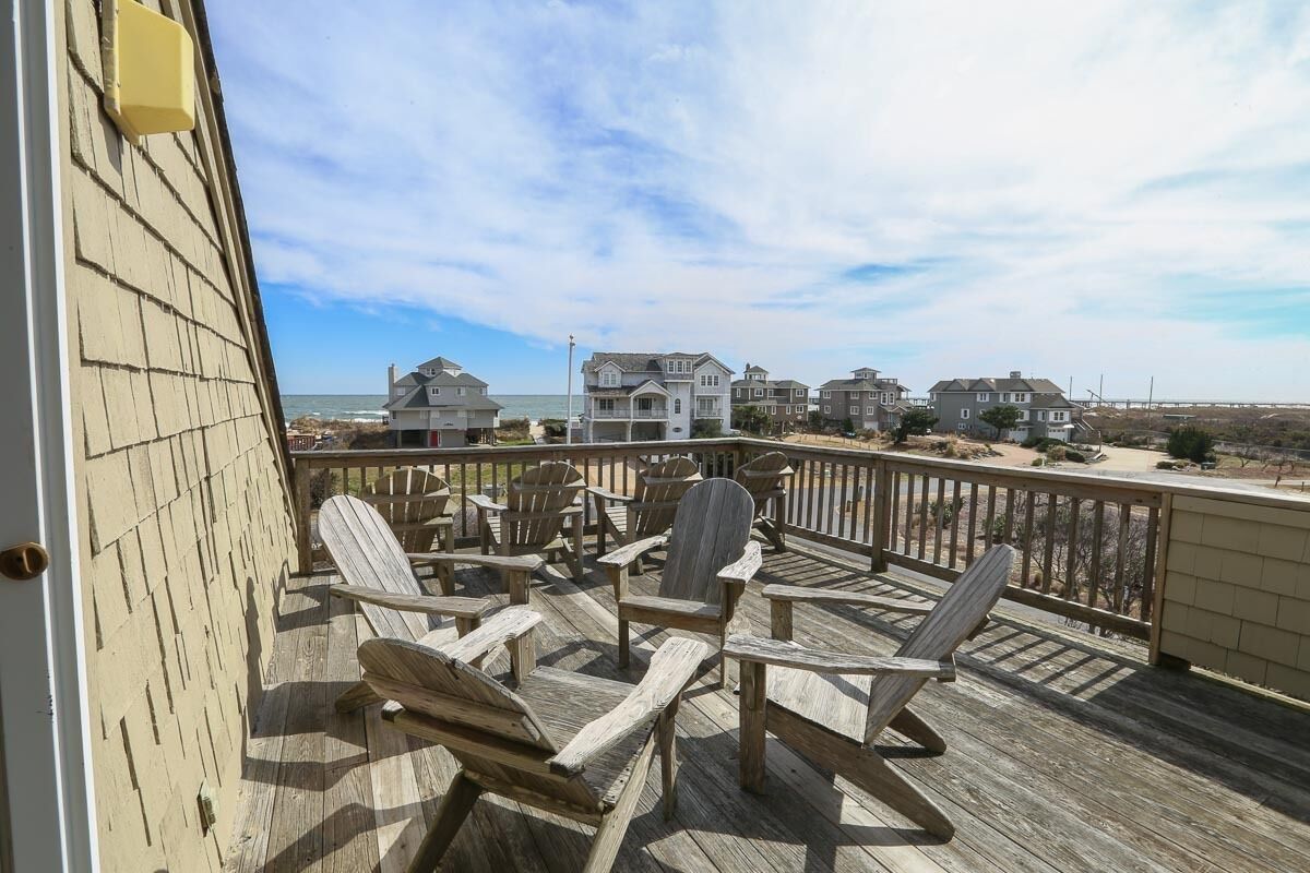 Eine Holzterrasse mit Adirondack-Stühlen mit Blick auf einen Strand und Häuser.