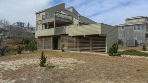 A modern two-story house with a balcony on the upper floor, a garage, and a small front yard with sparse vegetation. - Knock On Wood - Directly across from the beach access with terrific ocean views (Duck)
