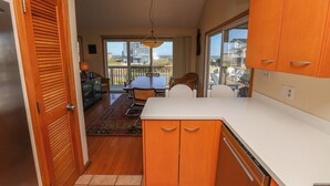 A compact kitchen with wooden cabinets and a white countertop, leading to a dining area with a table and chairs. - Knock On Wood - Directly across from the beach access with terrific ocean views (Duck)