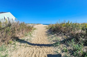 A sandy path leading to a beach, flanked by vegetation and a building with a light blue exterior. - Captain's Cove - Watch the ocean waves from your front porch! (Kitty Hawk)