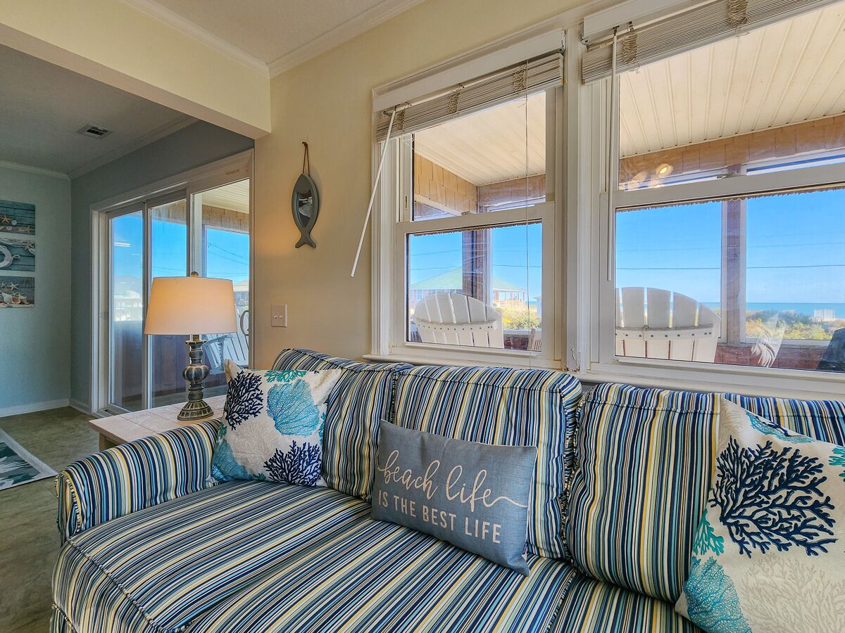 Ein gestreiftes Sofa mit einem Kissen mit der Aufschrift 'beach life is the best life', einer Lampe und Blick auf den Ozean durch große Fenster.