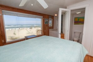 A bedroom with a large window overlooking the beach, a bed, a chair, and a bathroom with a toilet and a painting. - RETREAT - Charming and cozy 3 BR one level classic beach cottage! (Kitty Hawk)