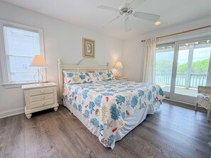 Quarto com cama, mesas de cabeceira, ventilador de teto, janela com persianas e vista para uma varanda.