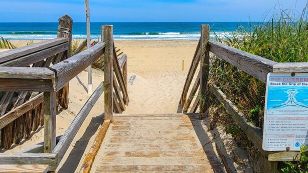 Une promenade en bois mène à une plage de sable, et un panneau indique la présence de courants de contre-rivage dangereux.