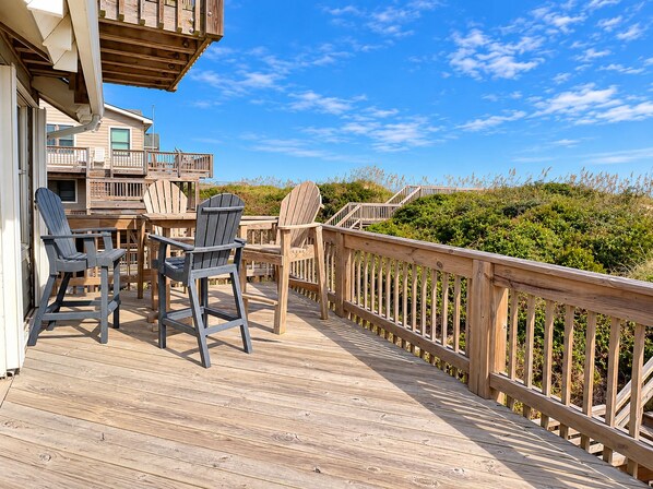 A wooden deck with two Adirondack chairs, a wooden railing, and a view of the beach and vegetation.