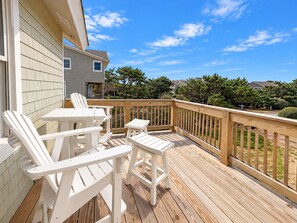 Una terraza de madera con sillas y mesas blancas, con vistas a una zona residencial con casas y árboles.