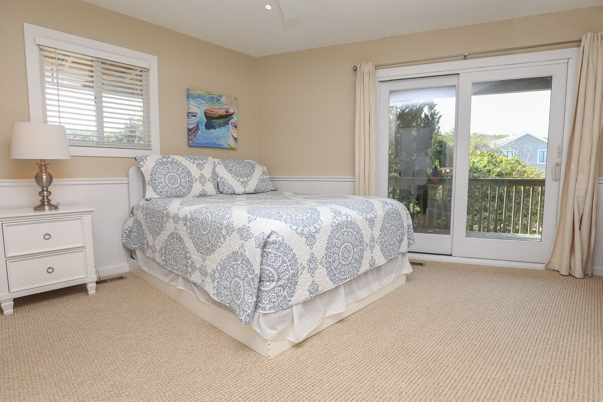 A bedroom with a bed, a nightstand, a window with blinds, a painting, and a sliding glass door to a balcony.