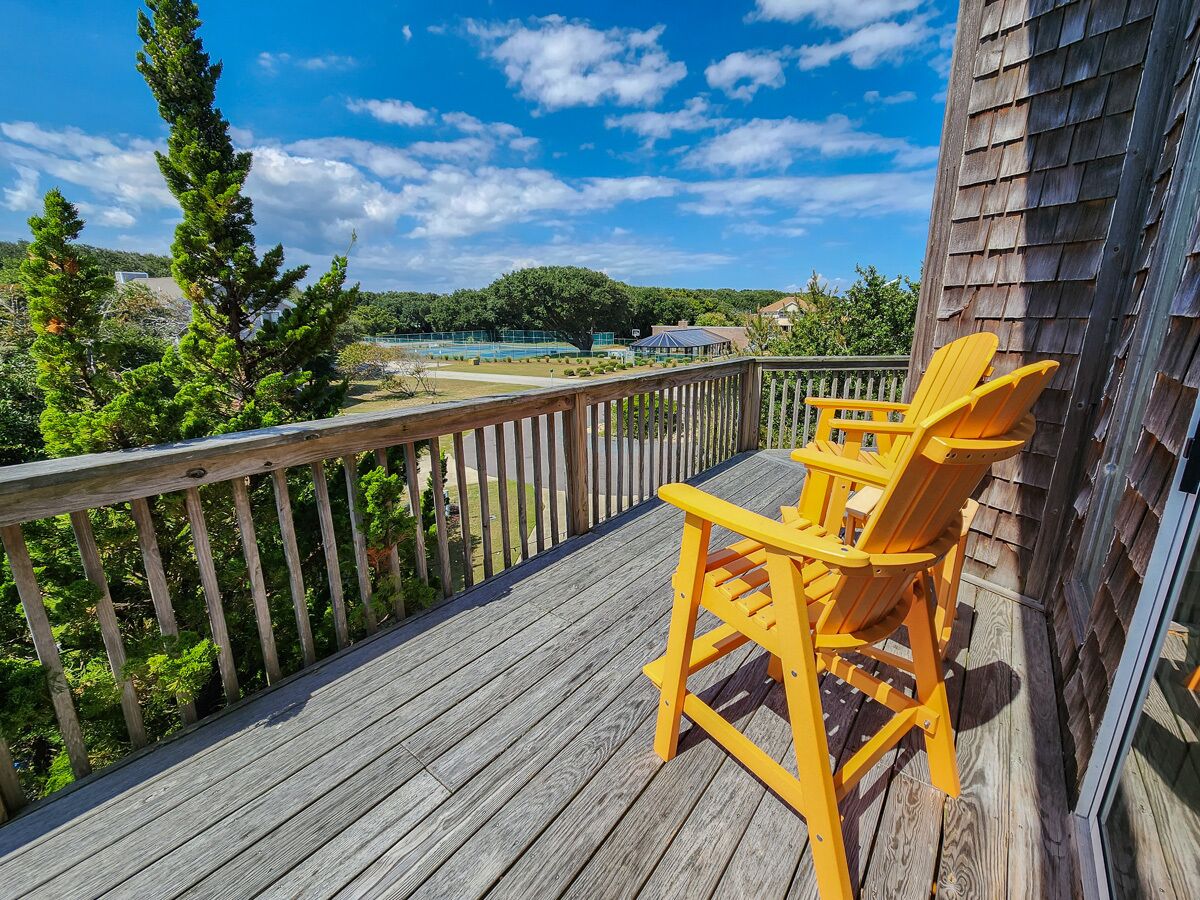 Une terrasse en bois avec un fauteuil Adirondack jaune, donnant sur un espace clos avec des arbres et un bâtiment en arrière-plan.