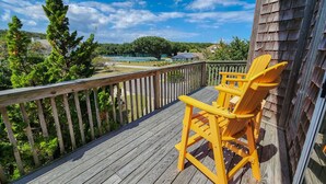 A wooden deck with a yellow Adirondack chair, overlooking a fenced area with trees and a building in the background. - “Surf & Sound” is the best of both worlds! Just steps away from the beach and Sound! (Duck)