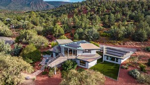 A modern two-story house with solar panels, surrounded by a landscaped garden and a view of a forested hillside. - Red Rock-view Sedona retreat with 2 bedrooms, 2 baths, and fireplace. (Sedona)