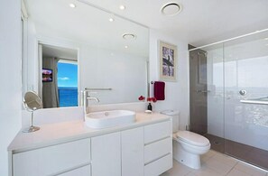 Un baño moderno con un espejo grande, lavamanos blanco y ducha con mampara de vidrio.
