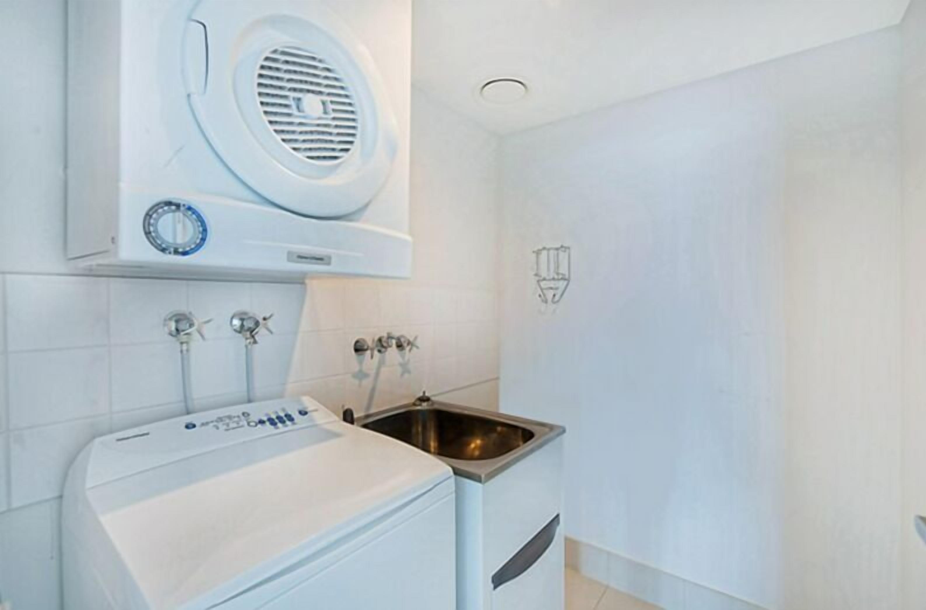 Ein kleines Badezimmer mit einem weißen Waschbecken, einer eingebauten Waschmaschine und einer Wandlüftungseinheit.