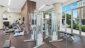 Un gimnasio moderno con diversas máquinas de ejercicio, un televisor en la pared y amplios ventanales con vistas a la ciudad.