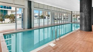 Una piscina interior con paredes de vidrio transparente, suelo de baldosas y vistas a los edificios exteriores.