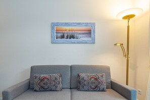 A grey sofa with patterned cushions, a framed painting of a sunset, and a floor lamp.