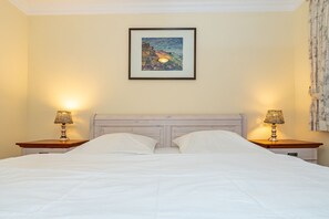 Een bed met witte lakens, twee nachtlampjes en een lijst met een schilderij aan de muur.