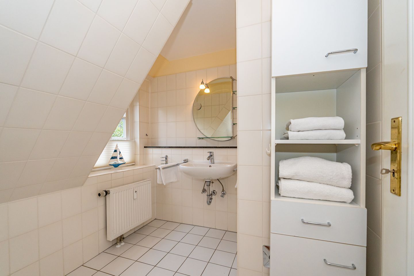 Une salle de bain moderne avec un lavabo blanc, un miroir et un porte-serviettes. Il y a un radiateur sous le lavabo et une fenêtre donnant sur l’extérieur.