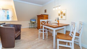 Ein Essbereich mit Holztisch und weißen Stühlen, einer braunen Ledersofa, einem Holzschrank und einem Fernseher auf einem Glastisch.