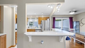 Una cocina moderna con encimeras blancas, armarios de madera y electrodomésticos de acero inoxidable. Cuenta con un comedor con mesa y sillas de vidrio, y vistas al exterior a través de amplias ventanas.