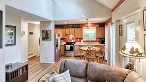 A spacious living room with a fireplace, a dining area with a round table and chairs, and a kitchen with wooden cabinets and a window.
