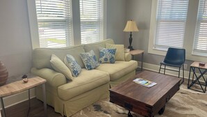 Ein Wohnzimmer mit einem beigefarbenen Sofa, blauen Kissen mit Muster, einem Holztisch, einem Beistelltisch, einer Lampe und einem blauen Sessel.