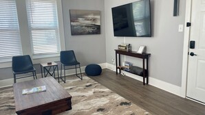 Ein modernes Wohnzimmer mit einem Holztisch, zwei Stühlen, einem blauen Hocker, einem an der Wand montierten Fernseher und einem Konsolentisch mit Teppich.