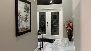 A hallway with a framed painting of palm trees, a door with decorative glass panels, a black table with a small item on it, and a vase with flowers.