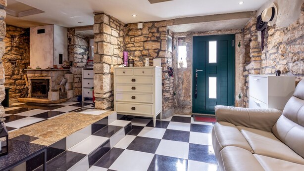 Un salon avec un sol à carreaux, un mur en pierre, une cheminée, une commode blanche et une porte verte.