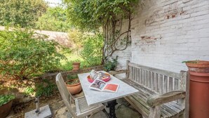 A wooden bench and table set against a white brick wall, surrounded by potted plants and greenery. - 29 Wentworth Road, Aldeburgh (Aldeburgh)