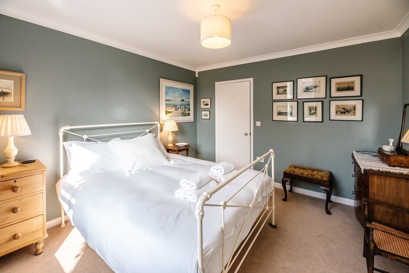 A bedroom with a metal bed frame, a wooden nightstand, a bedside table with a lamp, a wooden bench, and a dresser.