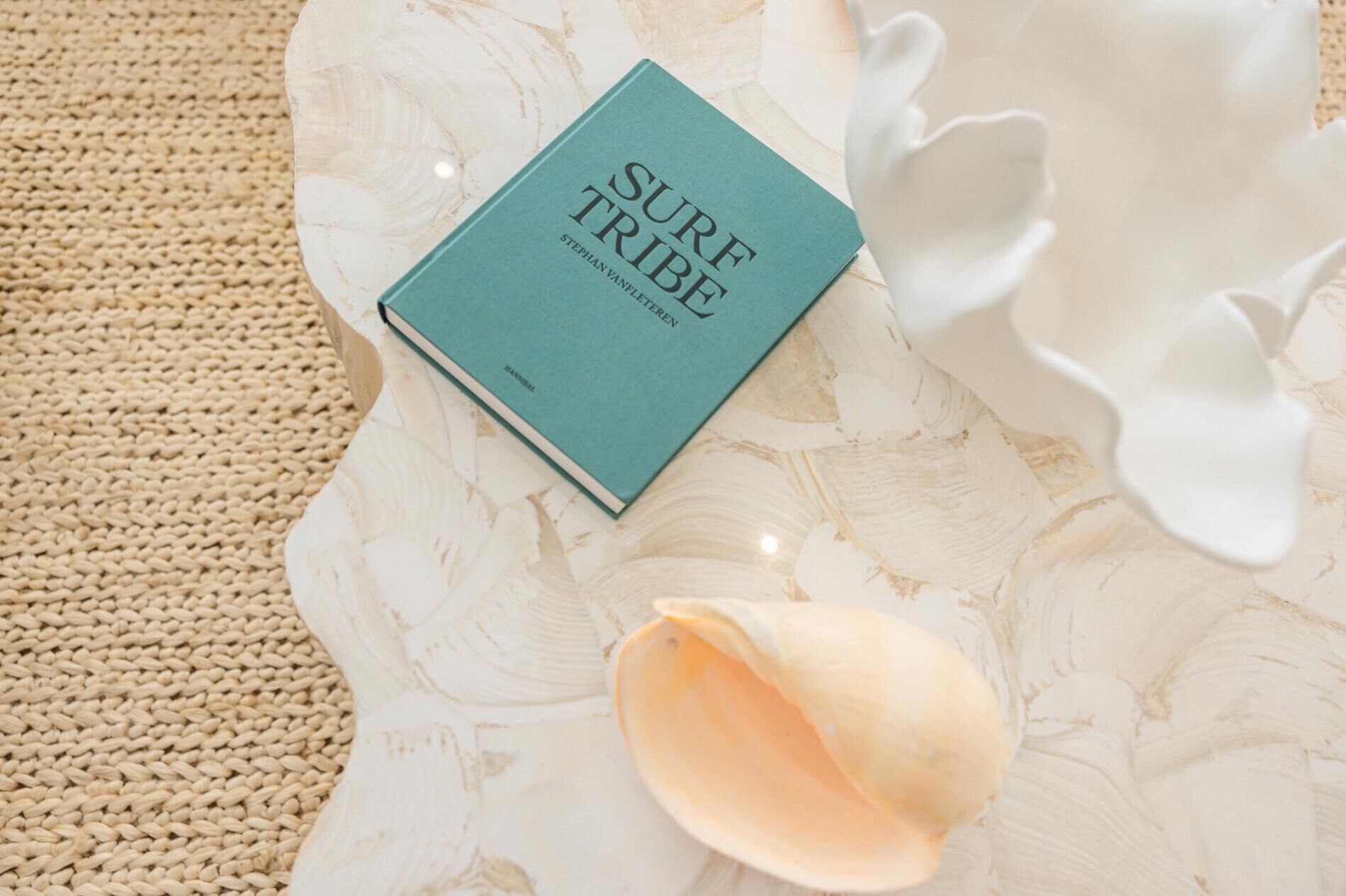 Een boek met de titel 'SURF TRIBE' op een structuurrijk oppervlak, met een geweven mat en een wit, organisch vormig object.