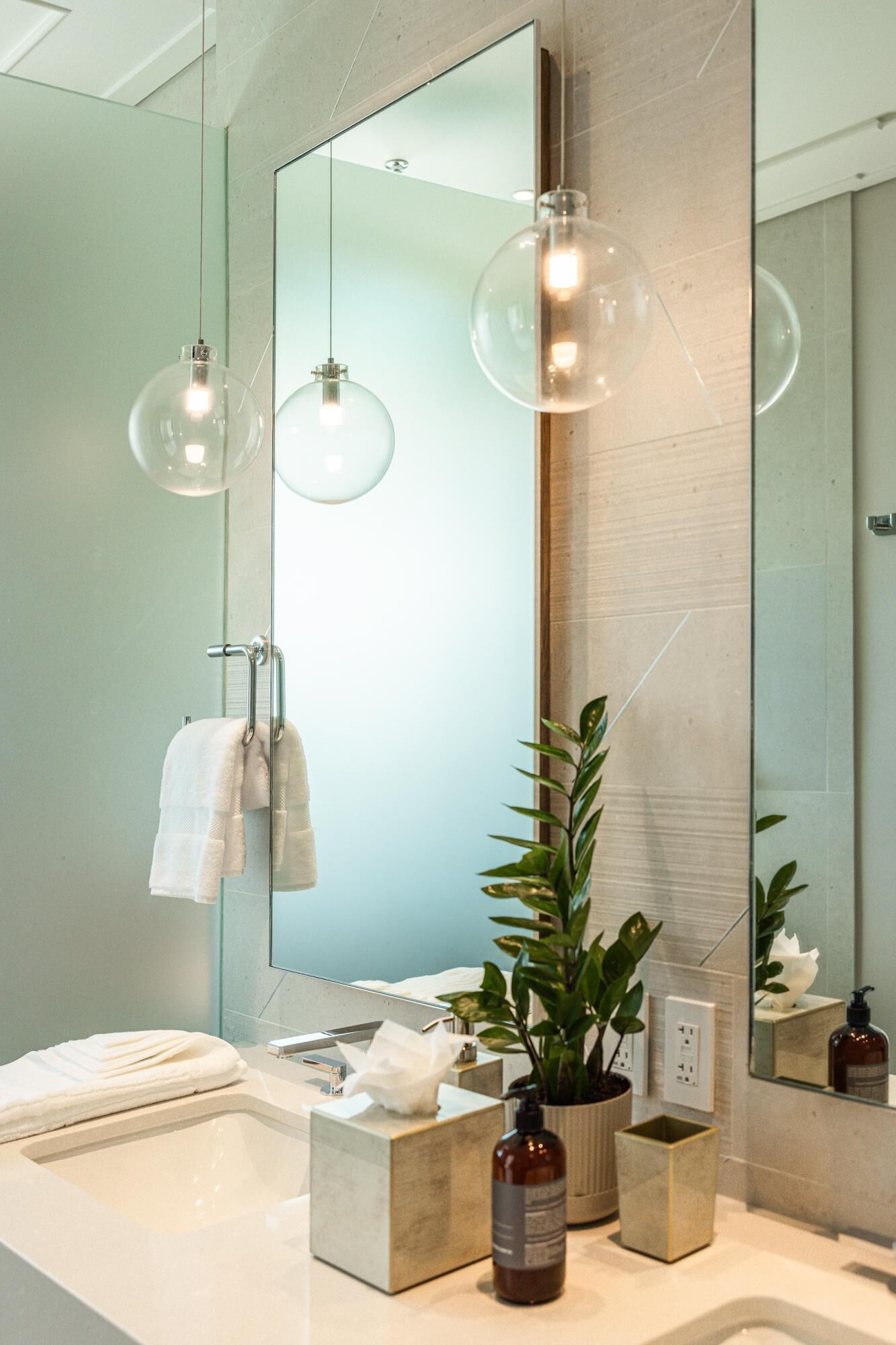 A modern bathroom with a large mirror, two hanging pendant lights, a white sink, a plant, and a towel rack.