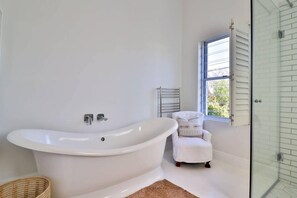 Un baño moderno con una bañera independiente, un sillón blanco pequeño, un box de ducha de vidrio y una ventana con persianas.