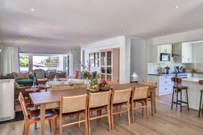 A spacious living area with a dining table, wicker chairs, a sofa, and a kitchen with white cabinets and a large window.