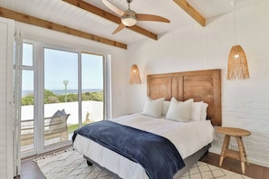 Quarto com uma cama grande, vigas de madeira no teto, um ventilador de teto e vista através de uma porta de correr de vidro.