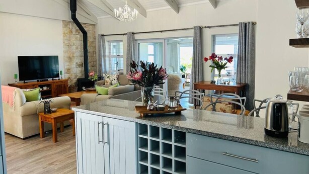 A modern kitchen with a central island, a dining area with a table and chairs, a living room with a sofa and TV, and a stone fireplace.
