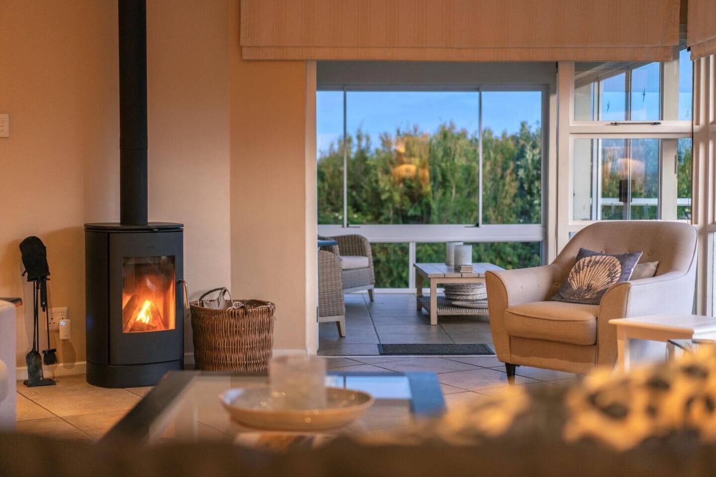 A cozy living room with a wood-burning stove, a comfortable sofa, and a view of greenery through large windows.