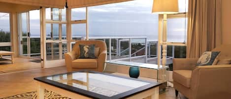 A modern living room with a glass coffee table, two sofas, a chair, and large windows offering a view.