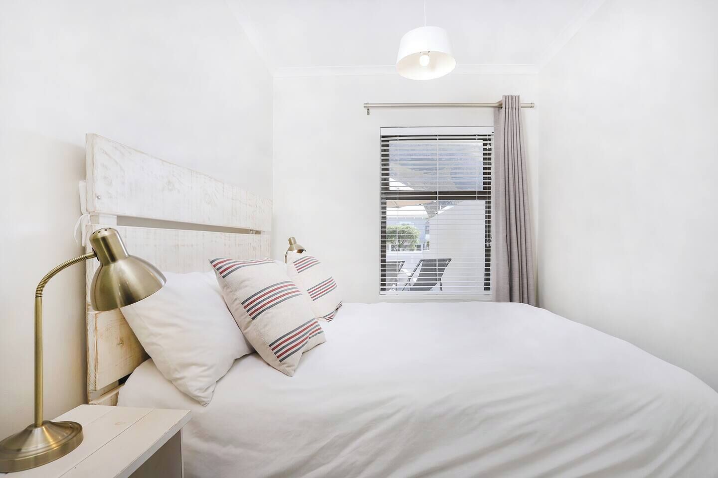 Un dormitorio con cama, lámpara de noche, ventana con persianas y vistas al exterior.
