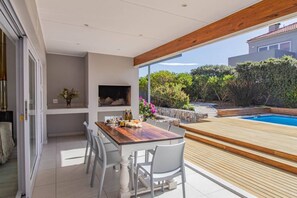 Um pátio moderno com mesa e cadeiras de madeira, TV embutida e vista para uma piscina e vegetação.