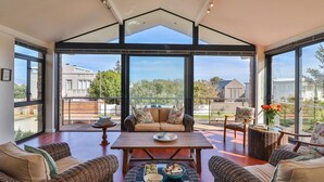 A spacious room with a high ceiling, large windows, a central seating area with a table, and wicker furniture. - Seaside Symphony Villa | Walking distance to Grotto Beach (Hermanus)