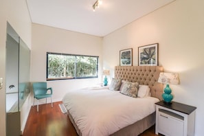 A bedroom with a large bed, a chair, a nightstand, and a window with a view of trees. - Seaside Symphony Villa | Walking distance to Grotto Beach (Hermanus)