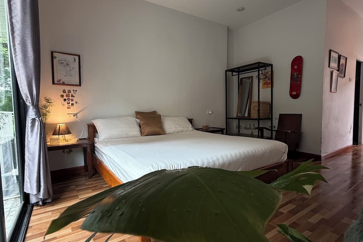 A bedroom with a bed, a nightstand, a lamp, a large green plant, and a window with curtains.
