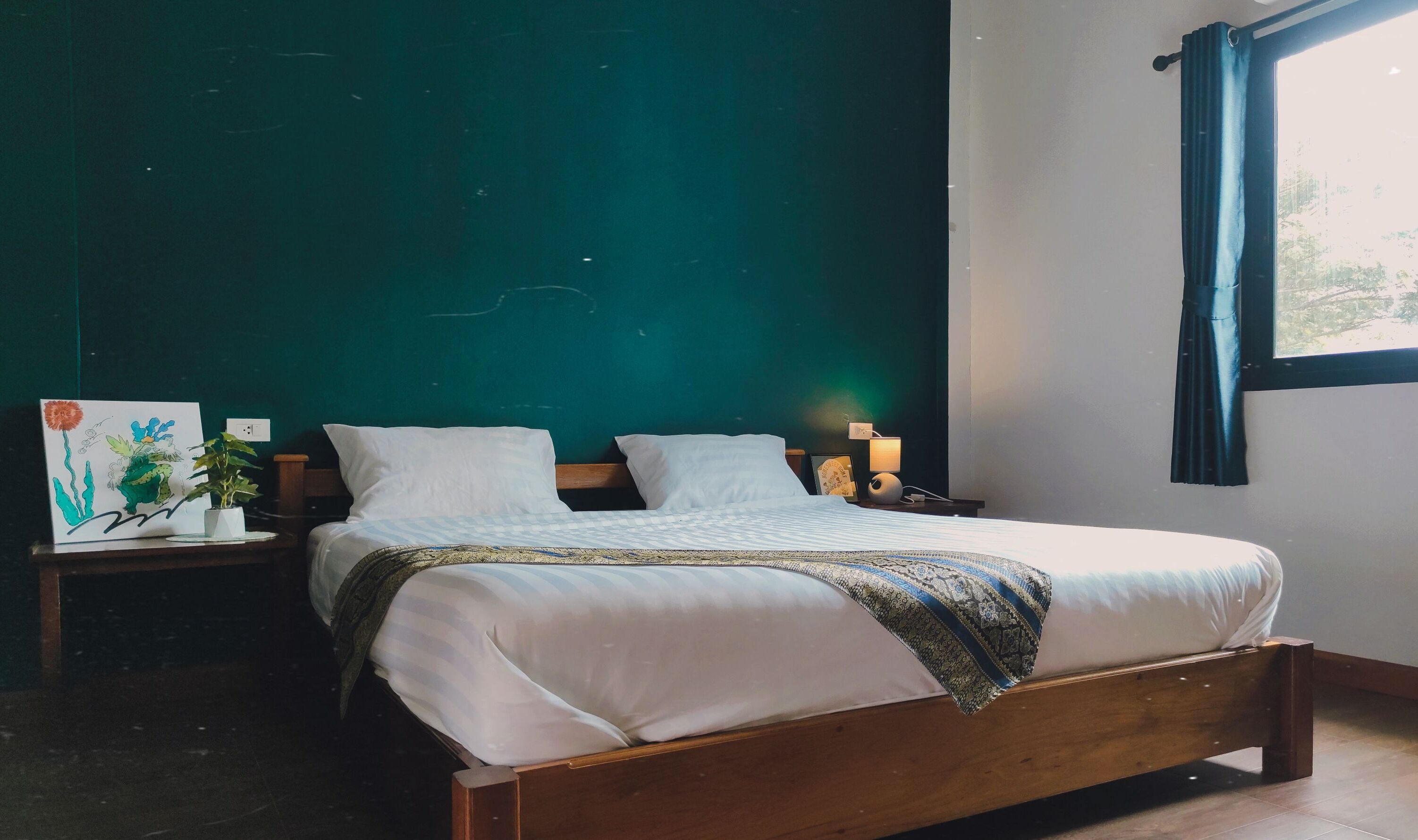 A neatly made bed with white linens and a patterned throw, a bedside table with a lamp, and a wall-mounted painting.